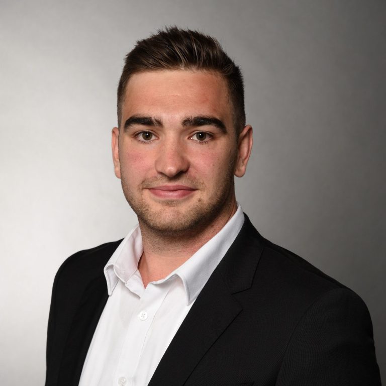 Ingenieurs-Portrait Robin Dietrich Junger Mann mit kurzen Haaren, Anzugjacke und Hemd vor einem grauen Hintergrund.