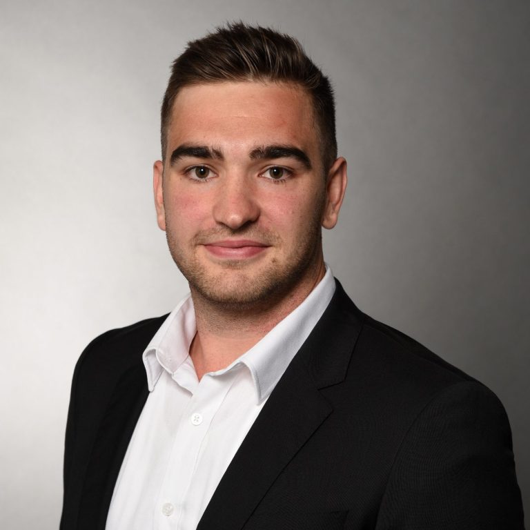 Junger Mann im Anzug mit weißem Hemd und kurzem, braunem Haar, neutraler Hintergrund.