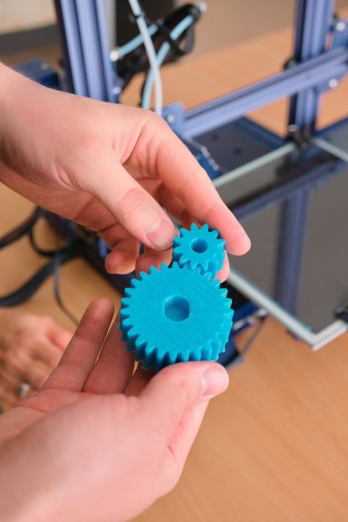 Zwei türkisfarbene Zahnräder werden von einer Person gehalten, im Hintergrund ein 3D-Drucker.