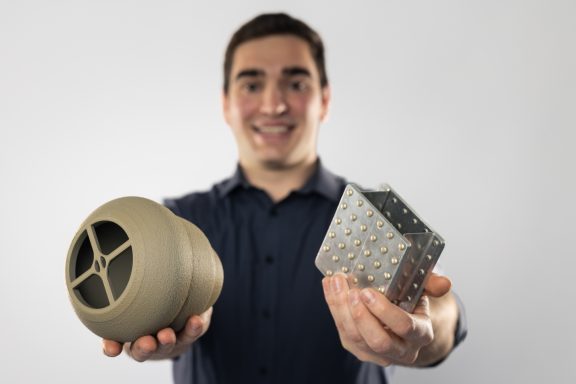 Mann hält zwei Objekte: einen runden, beige Behälter und einen würfelförmigen, silbernen Block.